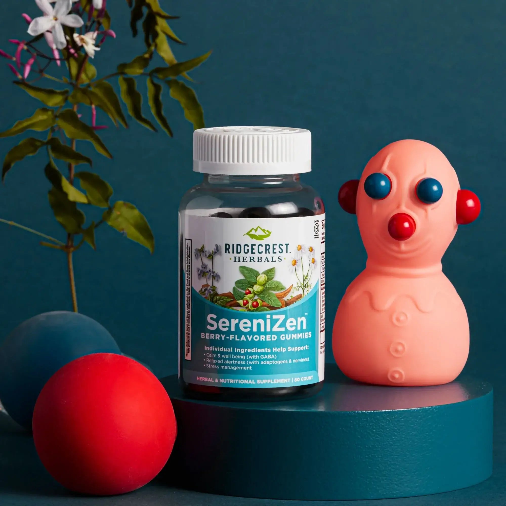 SereniZen Gummies bottle on a teal pedestal with a toy figure, colorful balls, and a flowered stem against a dark teal background